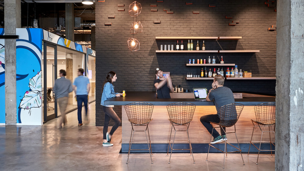 A group of people standing around a table with chairs and a person sitting at it.